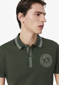 Ciemnozielona koszulka polo z teksturowanym materiałem, paskowanym kołnierzykiem, zapięciem na dwa guziki oraz haftowanym logo na piersi.