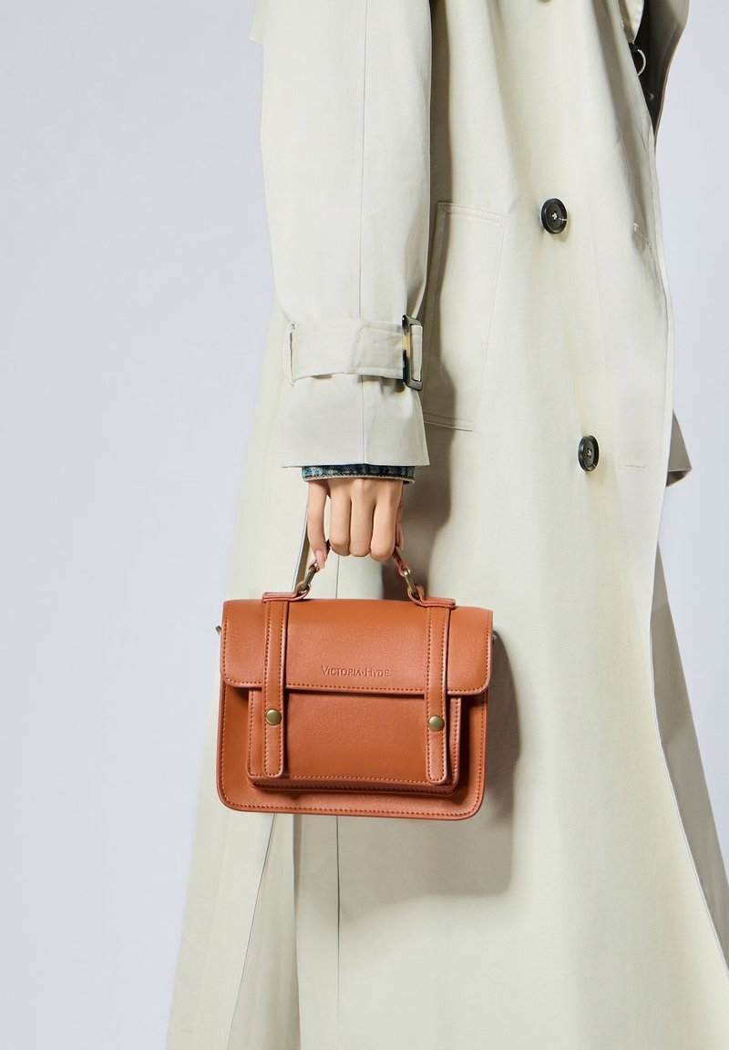 Main tenant un petit sac à main en cuir marron avec poche avant, porté sur un trench-coat beige avec de gros boutons.