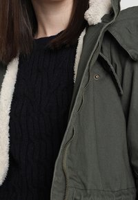 Olivově zelená parka s límcem podšitým fleecem, doplněná tmavě námořnicky modrým pleteným svetrem s copánkovým vzorem. Má zapínání na zip a boční kapsy.