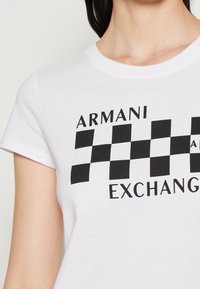 Bílý bavlněný tričko s černým kostkovaným vzorem a textem "ARMANI EXCHANGE" v tučném písmu. Krátké rukávy, kulatý výstřih.