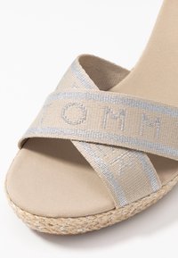 Beige sandaal met gekruiste stoffen bandjes met zilveren accenten, jute omhulde zool en een gladde, matte textieloppervlak.