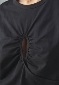 Gros plan sur une chemise noire avec une découpe ovale froncée au niveau de la poitrine, laissant apparaître une petite partie de peau en dessous.