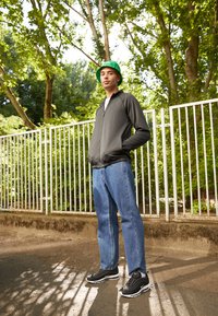 Giacca grigia con zip, t-shirt bianca, jeans blu, sneakers nere con suola bianca, cappello a secchiello verde, in piedi accanto a una recinzione bianca in un parco.