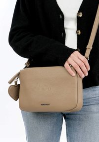Light brown leather crossbody bag with a textured finish, zip closure, and a heart-shaped charm attached. Adjustable strap.