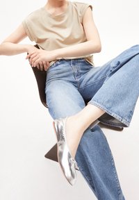 Zapatos planos plateados con un leve brillo, combinados con jeans azul de cintura alta y una blusa corta beige, sentada en una silla.