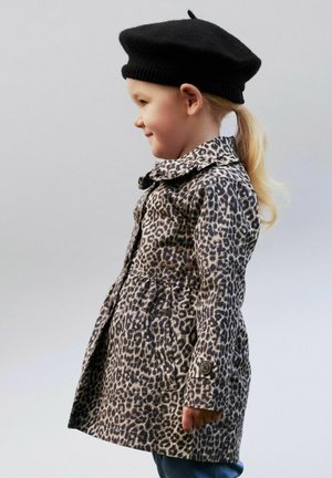 Noor laps külgprofiilis, kandes musta baretti ja nupudega ning taskutega leopardimustrilist mantlit heleda tausta ees.