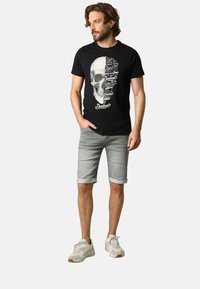 T-shirt en coton noir avec un graphisme de crâne et du texte sur le devant, associé à un short en denim gris clair et des baskets blanches avec des accents gris.