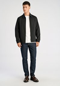 Czarna kurtka na zamek, pod spodem biała koszulka, niebieskie dżinsy, brązowe buty. Posiada kołnierz i zapięcie na zamek. Gładka tekstura, casualowy krój.