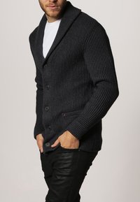 Mörkgrå, räfflad stickad kofta med sjalkrage, sex knappar och sidofickor. Bärs över en vit skjorta och matchas med svarta byxor.