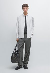 Camicia bianca a righe button-down sopra una t-shirt grigia, abbinata a pantaloni grigi. Borsa in pelle nera in mano, scarpe nere, sfondo minimalista.