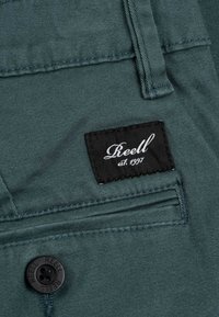 Pantalones de tela verde con una etiqueta negra cosida que lleva el texto blanco "Reell est. 1997." Un botón negro con el grabado "REELL" es visible.