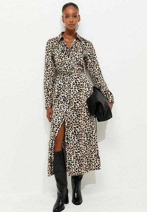 Mujer con vestido de manga larga con estampado de leopardo y abertura frontal, botas negras hasta la rodilla, pendientes de aro y llevando un bolso de mano negro.