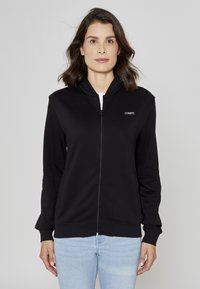 Sweat à capuche noir à fermeture éclair en matière douce avec un col haut, comportant un petit écusson logo sur la poitrine gauche et des poignets côtelés.