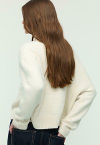 Cream-colored sweater with a textured, knitted design, ribbed hem, and loose sleeves; paired with dark denim pants, viewed from behind.