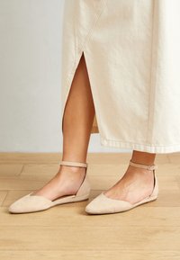 Ballerines en nubuck beige avec un bout pointu et une sangle à la cheville. Design minimaliste avec une semelle plate et des côtés ouverts. Sol en bois en arrière-plan.