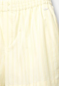 Short jaune clair avec des rayures blanches verticales, taille élastique, deux boutons, poche latérale et petite étiquette métallique de marque.