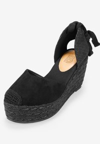 Black wedge sandal featuring a suede upper, woven jute platform, and a fabric ankle strap with bow detail. Closed toe with textured accents.