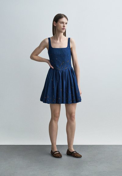 Γυναίκα όρθια με το χέρι στη μέση, φοράει αμάνικο μπλε φόρεμα με κεντήματα λουλουδιών και φλατ παπούτσια με λεοπάρ μοτίβο σε γκρι πάτωμα.