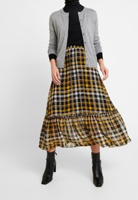 Grå knapp-up cardigan över en svart turtleneck, matchad med en rutig maxikjol i gult, svart och vitt, med rynkad nederkant. Svarta ankelskor.