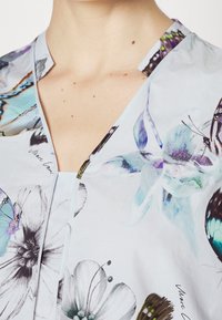 Floral printed blouse in light blue fabric. Features a V-neck, detailed butterfly and flower patterns, and subtle text accent. Smooth texture.
