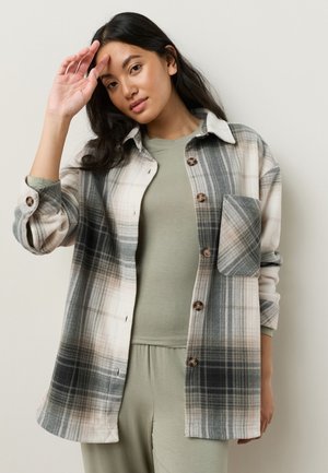 Joven con cabello largo y oscuro que lleva una camisa de cuadros beige y gris abotonada sobre una camiseta verde clara y pantalones, posando con una mano cerca de la frente.