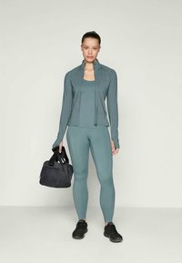 Fittes sportos öltözet türkiz színben, zipzáros dzseki és leggings tartozik hozzá. A modell egy fekete edzőtáskát tart. Sima textúra, könnyű anyag.