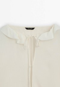 Blouse couleur crème avec un petit col à volants et un cordon à nouer au niveau de l'encolure, étiquetée Massimo Dutti.