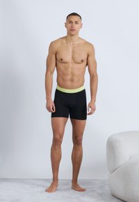 Αντικατοπτρίζοντας πάνω σε απλό φόντο, ένας άντρας χωρίς πουκάμισο στέκεται ξυπόλητος φορώντας μαύρα μποξεράκια με κίτρινη μέση, δίπλα σε μια λευκή καρέκλα με υφή.