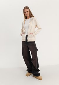 Felpa con zip beige dal finish testurizzato, con due tasche frontali, abbinata a jeans neri larghi e zoccoli color sabbia su uno sfondo neutro.