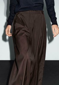 Pantalons plissés marron en tissu lisse, avec une taille cintrée et des poches, associés à un pull sombre texturé.