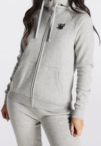 Lichtgrijze hoodie met ritssluiting, gemaakt van zacht materiaal, met een voorzak, trekkoordjes en een zwart logo geborduurd op de borst.
