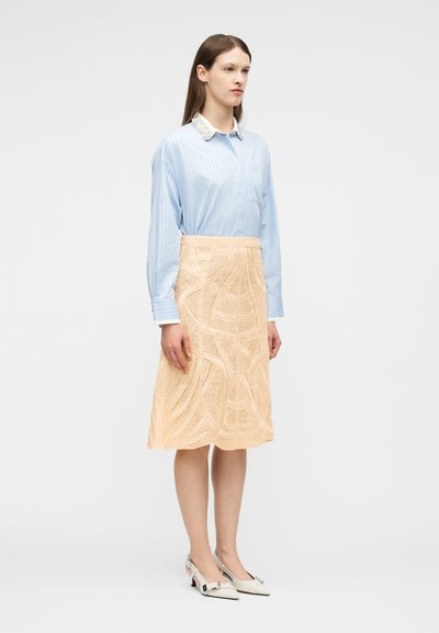 Une femme se tient debout, portant une chemise à rayures bleues avec un col en dentelle blanche, une jupe beige texturée jusqu'aux genoux et des chaussures blanches à petits talons avec des boucles.