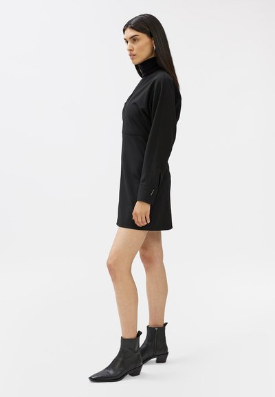 Rochie neagră cu mânecă lungă, gât înalt, cu un design ajustat, fante laterale și cusături minime, asortată cu botine negre până la gleznă.