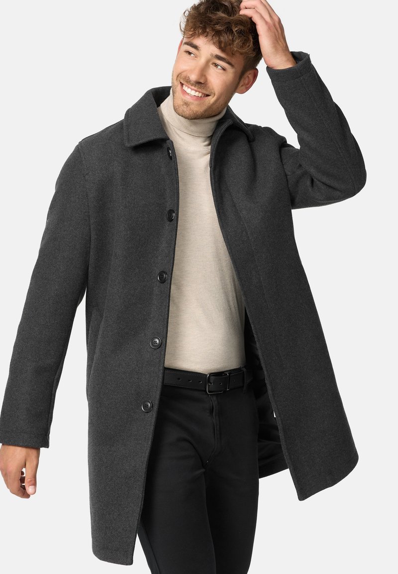 Grå uldfrakke med krave, fem sorte knapper og lige snit, båret over en lys beige turtleneck og sorte bukser.