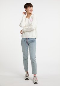 Witte trui met een V-hals en lange mouwen, met een roze hartaccent. Gecombineerd met lichtblauwe jeans en multikleurige sneakers.