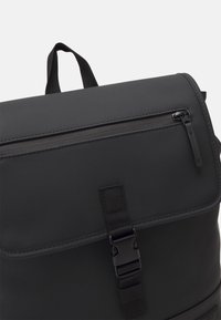 Zaino nero con finitura liscia e opaca, pattina superiore, tasca frontale con zip e tracolla regolabile con fibbia in plastica.