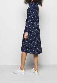 Nainen, joka on pukeutunut tummansiniseen pitkähihamaiseen mekkoon, jossa on valkoisia timanttikuvioita, yhdistettynä valkoisiin tennareihin, joissa on kultaiset kantakohdat, seisoo sivuttain.