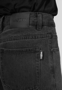 Svarta denimjeans med en texturerad yta, med en svart läderetikett i midjan och en sidetikett. Sömmen är synlig längs fickorna.