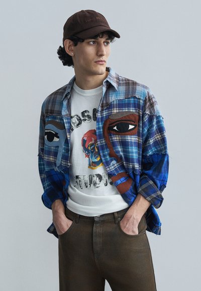 Πουκάμισο φανέλας καρό σε μπλε και καφέ με σχέδια προσώπων, φορεμένο πάνω από ένα λευκό γραφικό t-shirt. Καφέ καπέλο και σκούρα καφέ τζιν ολοκληρώνουν την εμφάνιση.