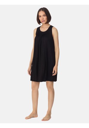 Vrouw met kort bruin haar, gekleed in een mouwloze zwarte jurk tot aan de knieën met geplooide halslijn, zonder schoenen staand tegen een effen achtergrond.