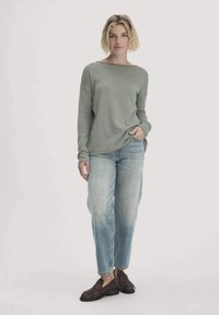 Langärmliges Oberteil mit Rundhalsausschnitt aus hellgrünem Stoff, kombiniert mit locker sitzenden, ausgewaschenen blauen Jeans und dunkelbrownem Slipper.