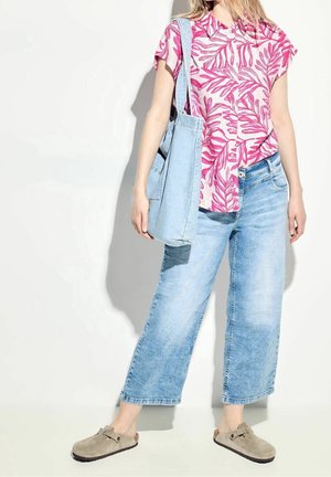 Kobieta w różowej koszuli w liście, jasnoniebieskie skrócone jeansy, beżowe buty wsuwane i nosząca jasnoniebieską torbę na ramię.