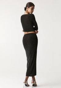 Čierny, trblietavý dvojdielny outfit obsahujúci top s dlhými rukávmi a priliehavú maxi sukňu s ruching detailami, doplnený o metalické sandále na tenkých podpätkoch.