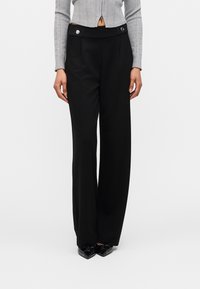 Femme portant un pantalon noir taille haute à jambes larges avec des détails boutons et des chaussures noires pointues, associé à un top gris en maille zippé.