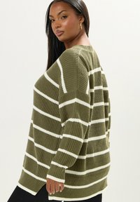 Grüner Strickpullover mit weißen horizontalen Streifen, lockerer Passform, langen Ärmeln und seitlichen Schlitzen am Saum. Strukturierter Stoff.
