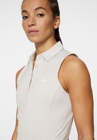 Ärm-lös polo skjorta i ljusbeige tyg, med krage, framplacket med knappar och en vit logotyp på bröstet. Slät textur.