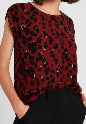 Kvinde iført en ærmeløs rød bluse med sorte og beige leopardpletter, stoppet ned i sorte bukser, hænderne i lommerne.