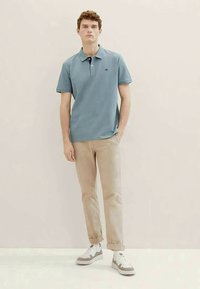 Gaiši zils polo krekls ar melnu apkakli, bēšas bikses un pelēkas sporta kurpes. Krekliņam kreisajā krūtīs ir neliels logo.