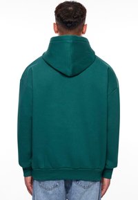Sweatshirt à capuche vert surdimensionné avec une capuche arrondie, des poignets et une taille côtelés, fabriqué à partir d'un tissu doux et présentant une texture lisse.
