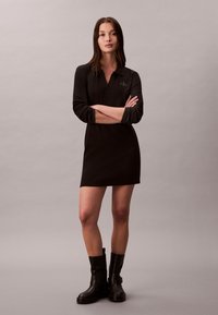 Femme debout, bras croisés, portant une robe polo noire à manches longues et des bottines noires, sur un fond clair uni.